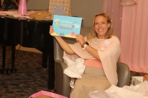 A smiling woman sits in a chair holding up a children's book titled "Letters, Numbers & Tillie Wren" during a Baby Shower Celebration. Gift bags are visible in the background.