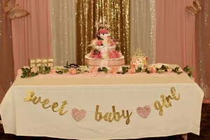 A decorated table with a "sweet baby girl" banner, a floral cake, roses, and charming pink and gold Baby Shower Celebration decorations.