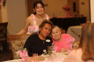 A young woman stands behind two older women seated at a decorated table with pink flowers, all smiling at the camera during a joyful Baby Shower Celebration in an indoor setting.