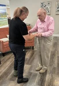 Ken, an elderly man in a pink shirt, holds hands with a woman in a black shirt as they stand on a wood floor in a living room setting, ready for some ballroom dancing.