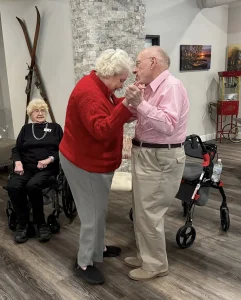 An elderly couple enjoys ballroom dancing in a room while Ken, seated in a wheelchair, watches and smiles; a walker and popcorn machine are visible in the background.