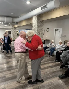 An elderly man named Ken and a woman enjoy ballroom dancing together in a common room as other seniors watch from chairs along the wall.