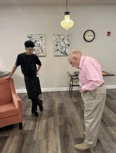 Ken, wearing a pink shirt and khaki pants, stands and looks down while a young man in black work attire leans against a red chair. A clock and framed art—perhaps hinting at ballroom dancing—hang on the wall behind them.