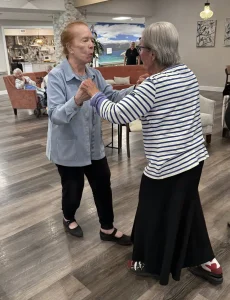 Two elderly women hold hands and dance together in a spacious room with wooden floors, gracefully enjoying ballroom dancing as several people, including Ken, watch in the background.