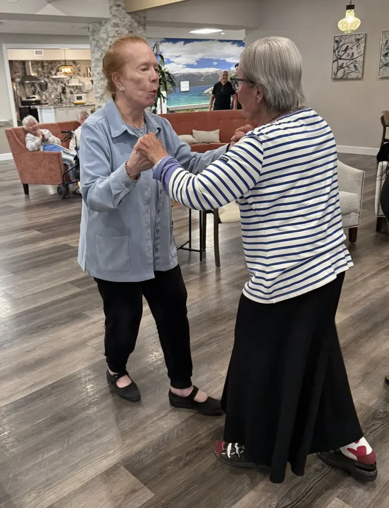 Two elderly women hold hands and dance together in a spacious room with wooden floors, gracefully enjoying ballroom dancing as several people, including Ken, watch in the background.