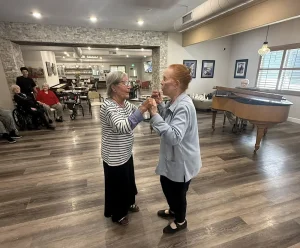 Two elderly women enjoy ballroom dancing together in a spacious room with wood flooring; other seniors, including Ken, are seated in the background near a piano.