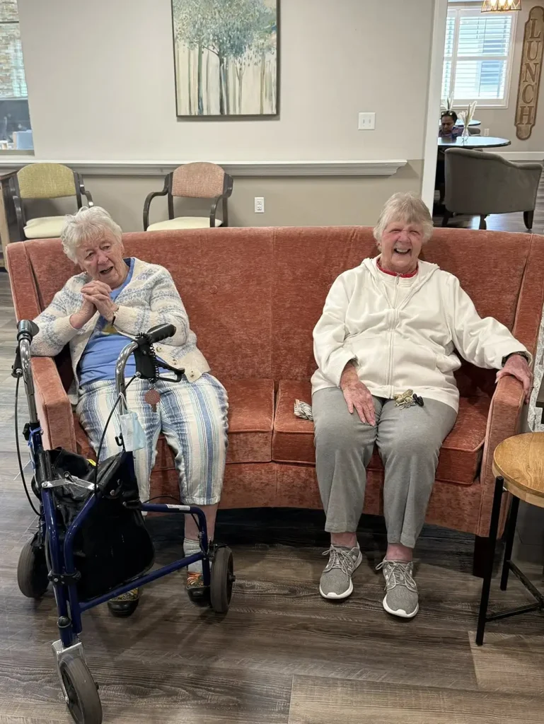 Two elderly women are sitting and smiling on a pink couch; one has a walker, and both are dressed casually in a room with neutral decor, reminiscing about their days of ballroom dancing with Ken.