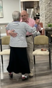 Ken and an elderly woman enjoy ballroom dancing together in a room with chairs and a stone accent wall, while another person sits in the background.