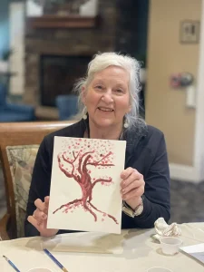 An older woman with gray hair smiles while holding up her art—a painting of a tree with pink blossoms—seated at a table indoors, proud of her delicate brushstrokes.