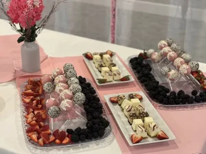 A dessert table with trays of cake pops, strawberries, blackberries, and assorted bite-sized cheesecake squares, decorated in a pink and white theme inspired by delicate blossoms and artful brushstrokes.