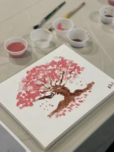 A canvas painting of a tree with pink blossoms sits on a table, surrounded by brushes and small cups of paint, the delicate brushstrokes capturing the essence of art and springtime beauty.
