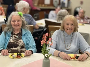 Two older women sit at a table with plates of dessert in front of them, smiling. A vase with blossoms adds charm to the scene, while other people are seated in the background.