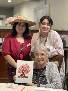 Three women pose indoors—one seated, holding a painting of pink blossoms, while two stand behind her, both smiling. Art supplies and brushstrokes fill the table, capturing their joyful creative moment.