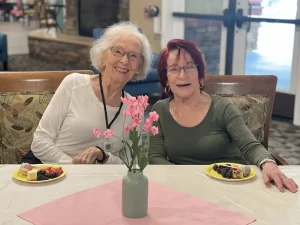 Two elderly women sit side by side at a table with desserts on yellow plates, a vase of pink blossoms in the center, smiling at the camera.