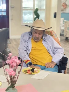 An elderly man in a wheelchair wearing a hat and yellow shirt sits at a table, surrounded by blossoms and fruit, the scene captured with gentle brushstrokes reminiscent of fine art.