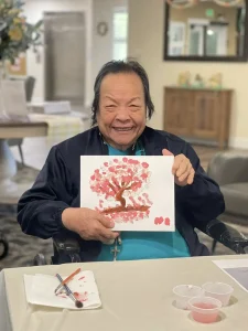 An older adult smiles while holding up a painting of pink blossoms. Art supplies, brushes, and paint cups are on the table, capturing the joy of creative brushstrokes.