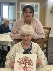 An elderly woman sits holding a painting of a tree adorned with blossoms, while a younger woman stands behind her. Both smile at the camera, sharing a joyful moment in the communal room filled with art.