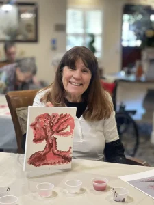 A woman smiles while holding up a painting of a red tree with beautiful blossoms; paint cups and art supplies are on the table in front of her.
