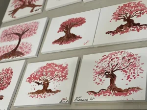 Several paintings of cherry blossom trees with delicate pink blossoms and expressive brushstrokes are displayed on a wall, each work of art signed by different artists.