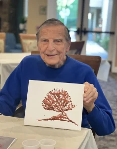 An elderly man in a blue sweater sits at a table, smiling and holding up his artwork—a painting of a tree with expressive brushstrokes on a white canvas, capturing delicate blossoms in brown paint.