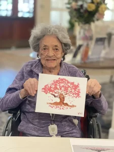 An elderly woman in a purple top sits in a wheelchair, holding up her art—a painting of a pink tree adorned with blossoms and delicate brushstrokes.