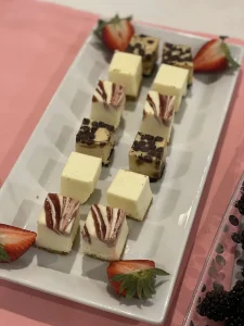 A rectangular white platter with three rows of assorted cheesecake bites, some topped with chocolate chips or fruit swirls, and garnished with halved strawberries—an artful display reminiscent of vibrant brushstrokes.
