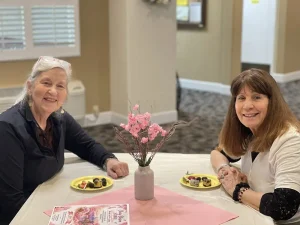 Two women sit at a table adorned with pink blossoms and plates of food, smiling at the camera in a brightly lit indoor setting.