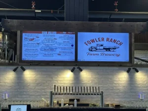A beer menu and Fowler Ranch Farm Brewery sign are displayed on screens above a bar with beer taps and glasses, set against a white tile wall—inviting guests to enjoy the atmosphere or start their Brewery Tour.