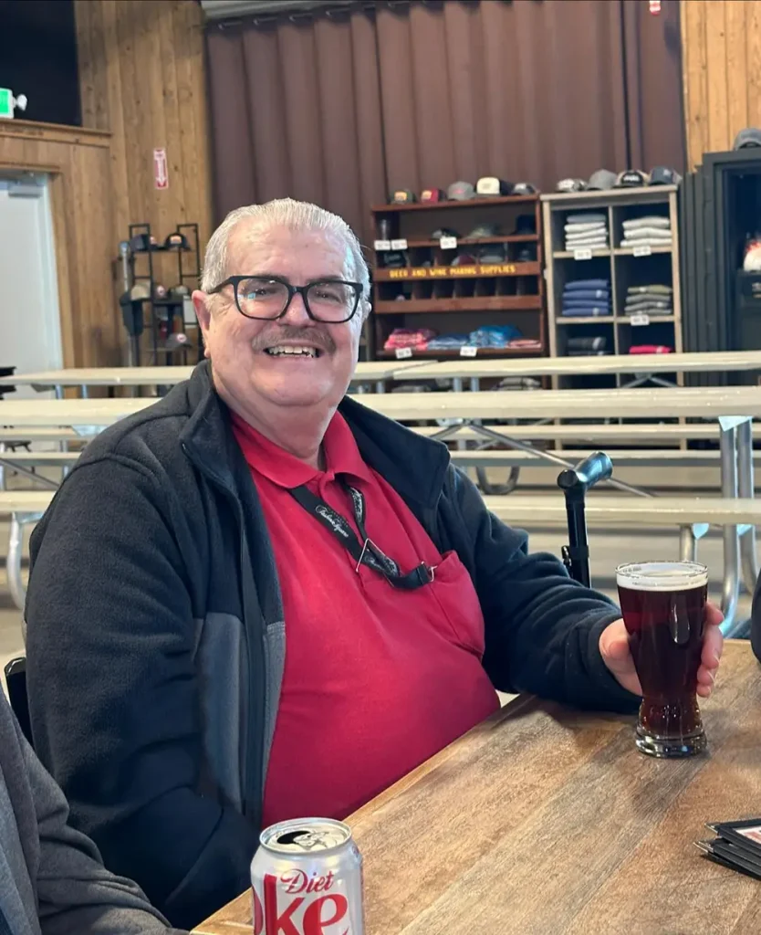An older man with glasses and gray hair sits at a table holding a dark beer, perhaps after a brewery tour. A can of Diet Coke, utensils, and shelves with folded shirts are visible in the background.