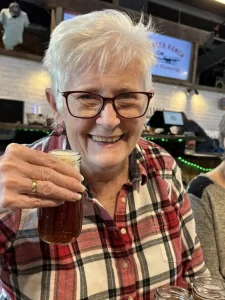 Older woman with short white hair, glasses, and a plaid shirt smiles while holding up a glass of beer during a brewery tour in a lively bar setting.