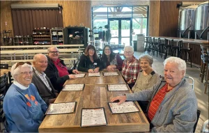 Eight people sit around a wooden table in a restaurant, each with a menu in front of them, eagerly discussing their upcoming brewery tour. The background shows empty tables and large windows.