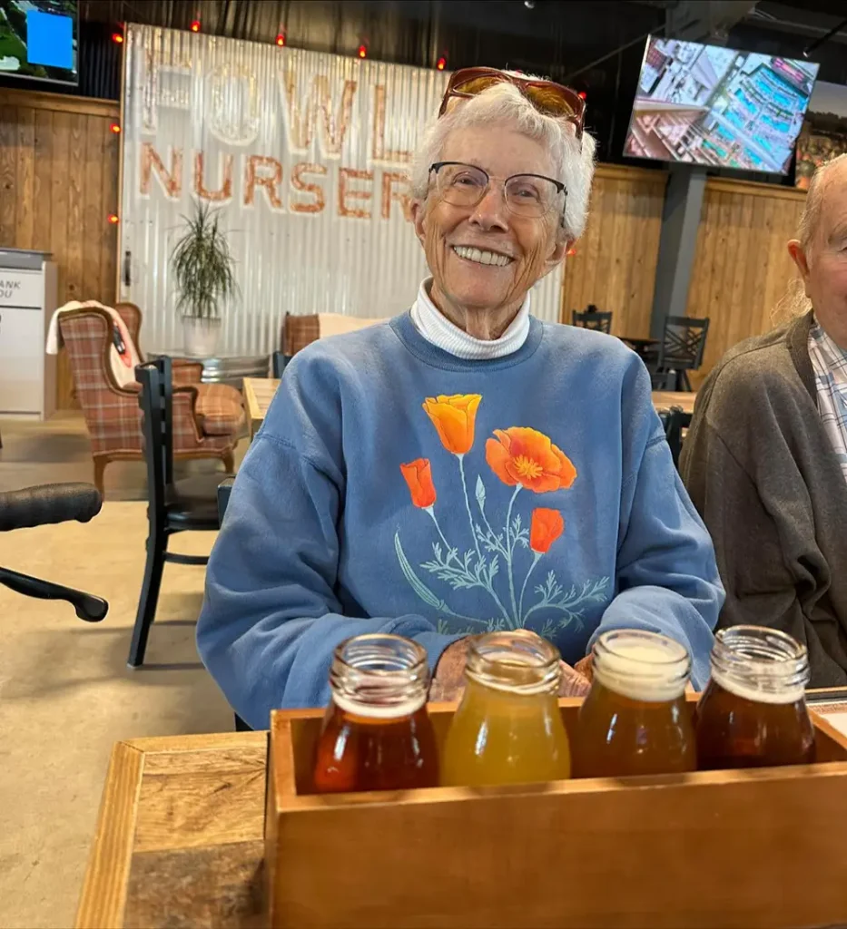 Elderly woman in a blue sweatshirt with a poppy design smiles at the camera during a brewery tour, a flight of four drinks in small glass bottles sits on a wooden tray in front of her.