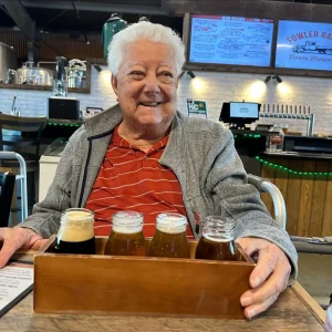 An older person smiles while sitting at a table in a brewery, holding a wooden tray with four small glasses for a beer tasting. Brewing equipment and a menu board add to the authentic brewery tour atmosphere in the background.