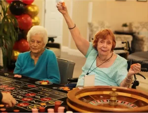 Two elderly women sit at a roulette table during casino night; one looks pleased and raises a chip in the air, while the other looks on quietly. Chips and roulette wheel are visible.