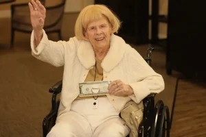 An elderly woman in a wheelchair waves and smiles, holding a novelty dollar bill. Dressed in white, she enjoys Casino Night festivities indoors.