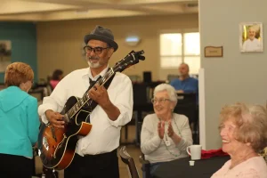 A man wearing a hat plays an electric guitar while elderly women sit at a table, smiling and clapping during Casino Night in a well-lit room.