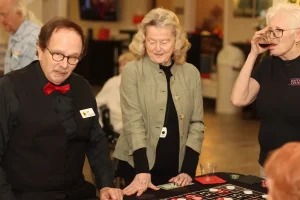 Three older adults gather around a Casino Night gaming table; one wears a dealer uniform with a red bow tie, while the others watch or sip drinks.