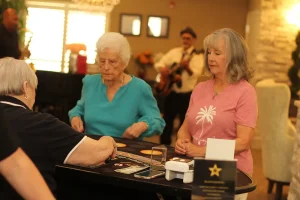 Three women sit at a table playing cards and chips during Casino Night, while a band performs in the background in a warmly lit indoor setting.