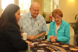 Three older adults stand around a gaming table with cards and chips, enjoying a lively Casino Night activity indoors.