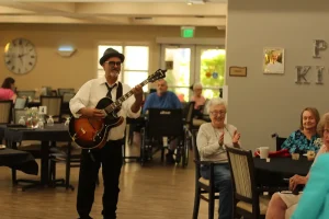 A man in a hat and sunglasses plays guitar while walking through a dining area during casino night, as elderly people seated at tables watch and clap.