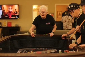 Two older women stand at a casino craps table during Casino Night, focused on the game. One holds chips, the other holds dice. A TV and other people are visible in the background.