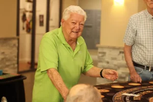 An older man in a light green polo shirt smiles while standing next to a table at Casino Night inside a well-lit room, with other people nearby.
