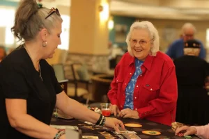 Two women stand at a table on Casino Night with cards and drinks; one woman in a red shirt smiles while the other focuses on the table.