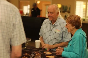 Two older adults stand at a casino game table with chips and a coffee cup, enjoying Casino Night as they interact with another person in a relaxed indoor setting.