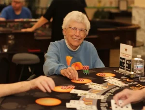 An older woman wearing glasses and a blue sweatshirt sits at a card table in a casino, smiling, with poker chips and playing cards in front of her.