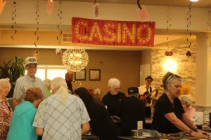 A group of seniors gather around casino game tables in a decorated room with a "CASINO" sign hanging above, enjoying an exciting Casino Night together.