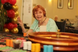 An elderly woman in a light blue top sits at a roulette table during Casino Night, smiling and holding a gaming chip, with colorful chips and a roulette wheel in front of her.