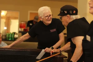 Two elderly women in black shirts talk by a pool table indoors during Casino Night; one woman holds a cue stick, while the other smiles and holds paper.
