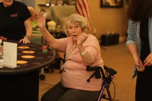 An elderly woman sitting on a walker plays poker at a Casino Night table, raising one hand and smiling, while others stand nearby handling poker chips.