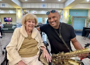 An elderly woman in a wheelchair sits next to a smiling man holding a saxophone inside a well-lit room decorated with small hanging flags, ready to enjoy the excitement of Casino Night.